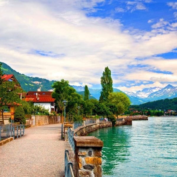 Scenic lakeside pathway with lush greenery, mountains in the background, and a clear blue sky, perfect for leisurely walks and nature photography.