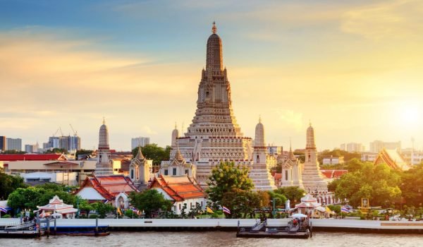 Wat Arun temple at sunset, showcasing intricate architecture and vibrant colors along the Chao Phraya River in Bangkok, Thailand. The scene includes lush greenery and traditional Thai structures, highlighting the cultural significance of this iconic landmark.