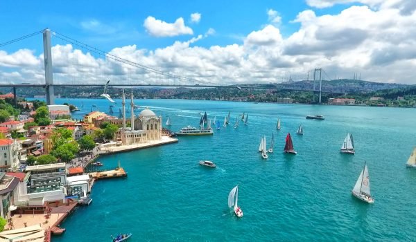 Aerial view of a vibrant waterfront scene featuring sailboats on a turquoise sea, with a historic building and lush greenery along the shore under a bright blue sky with fluffy clouds.