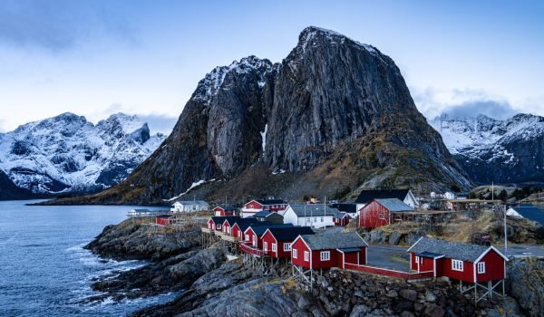 Scenic view of traditional red fishing cabins along the rocky coastline with dramatic mountains in the background, showcasing the natural beauty of Norway's landscape during twilight.