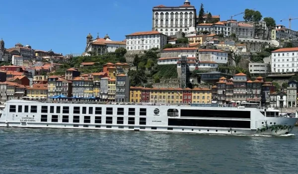 A scenic view of a modern river cruise ship docked along the picturesque waterfront of Porto, Portugal, with colorful hillside buildings and lush greenery in the background.