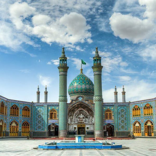 Imposing view of the stunning Sheikh Lotfollah Mosque in Isfahan, Iran, showcasing its intricate turquoise tiles, grand dome, and elegant minarets against a blue sky with fluffy clouds. The courtyard features a serene fountain surrounded by ornate architectural details.