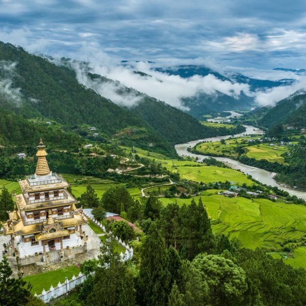 Aerial view of a Buddhist temple nestled in lush green mountains, overlooking a winding river and misty valleys in Bhutan.